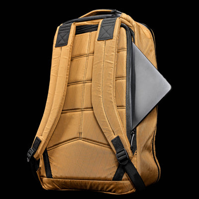 The GR1 USA - X-PAC is a tan, waterproof rucksack with black straps, showing a laptop partly visible in the side pocket against a black background.