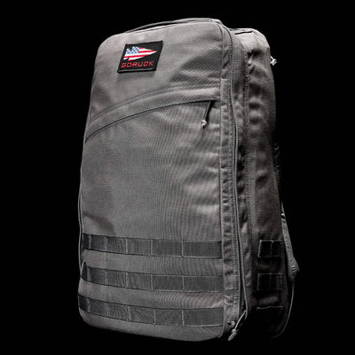 The GR1 USA - Cordura (The Original Ruck) by GORUCK is a gray backpack featuring a GORUCK patch, a front zippered pocket, and sturdy straps—masterfully crafted in American Manufacturing. Inspired by the resilience of Special Forces, this backpack stands proudly against a black background.