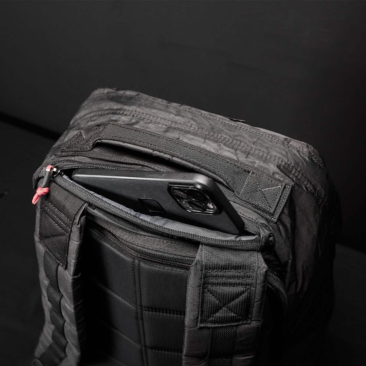 A close-up of the GR1 USA - Dyneema shows its sleek, black padded design. A smartphone in a matching black case peeks from a zippered compartment on the back, ensuring convenience. The durable construction by GORUCK is evident against a solid dark background.