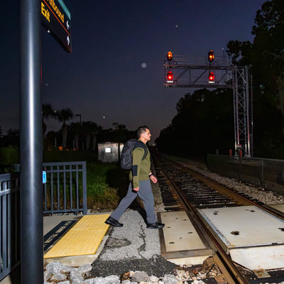 A person carrying a GORUCK GR1 USA - Dyneema rucksack crosses a railroad track at night. Red signal lights illuminate the tracks, with trees and an exit sign for the boardwalk visible in the background. The dark sky highlights the nighttime ambiance.