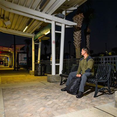 A man sits on a bench at a train station at night, clutching his GR1 USA - Dyneema rucksack from GORUCK. The station is illuminated with overhead lights, and palm trees are silhouetted in the background. The scene is quiet and empty, with only the rustle of leaves disrupting the stillness.