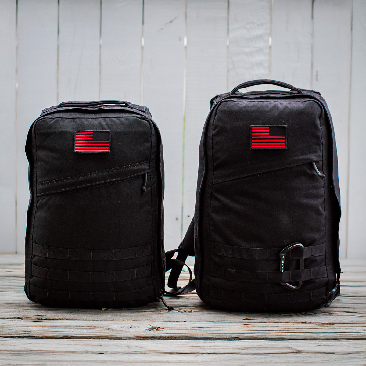 Two GR1 USA Throwback backpacks, black with red and black flag patches, sit on a wooden surface against a white fence. Made of rugged 1680 ballistic nylon for durability, they feature a bombproof laptop compartment for top-notch protection.