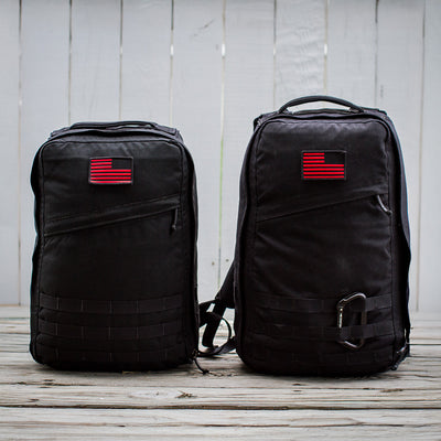 Two GR1 USA Throwback backpacks, black with red and black flag patches, sit on a wooden surface against a white fence. Made of rugged 1680 ballistic nylon for durability, they feature a bombproof laptop compartment for top-notch protection.