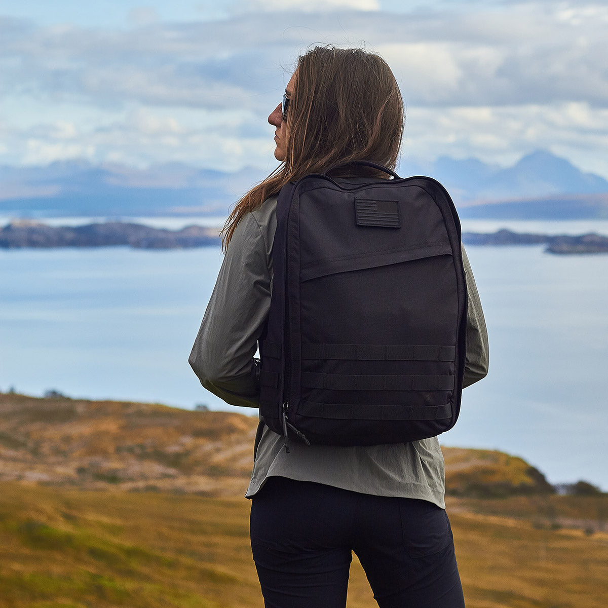 In an open landscape, a person with the GR1 USA Throwback backpack, made from durable 1680 Ballistic Nylon, stands ready for adventure. Sporting sunglasses and outdoor attire, they gaze at distant mountains with their gear secure in its bombproof laptop compartment.