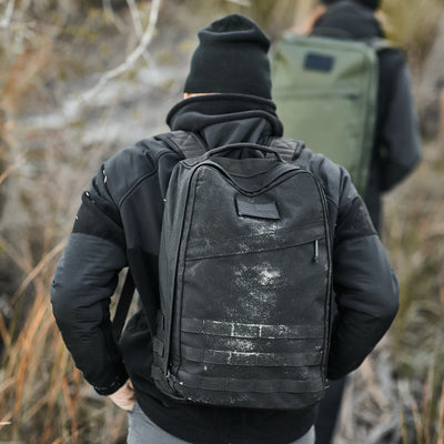 A person wearing a GR1 USA Throwback black backpack made of 1680 Ballistic Nylon walks outdoors with another person carrying a green backpack.