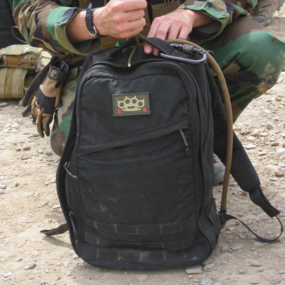 A person in a camouflage uniform stands on rocky ground, holding a black backpack with a brass knuckles patch. The pack features a bombproof laptop compartment made from durable 1680 ballistic nylon, similar to that of the GR1 USA Throwback gear.