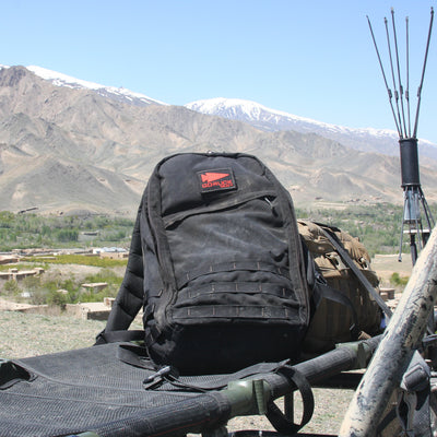 The GR1 USA Throwback, a black backpack made of 1680 Ballistic Nylon, rests on a cot with mountains silhouetted in the background.