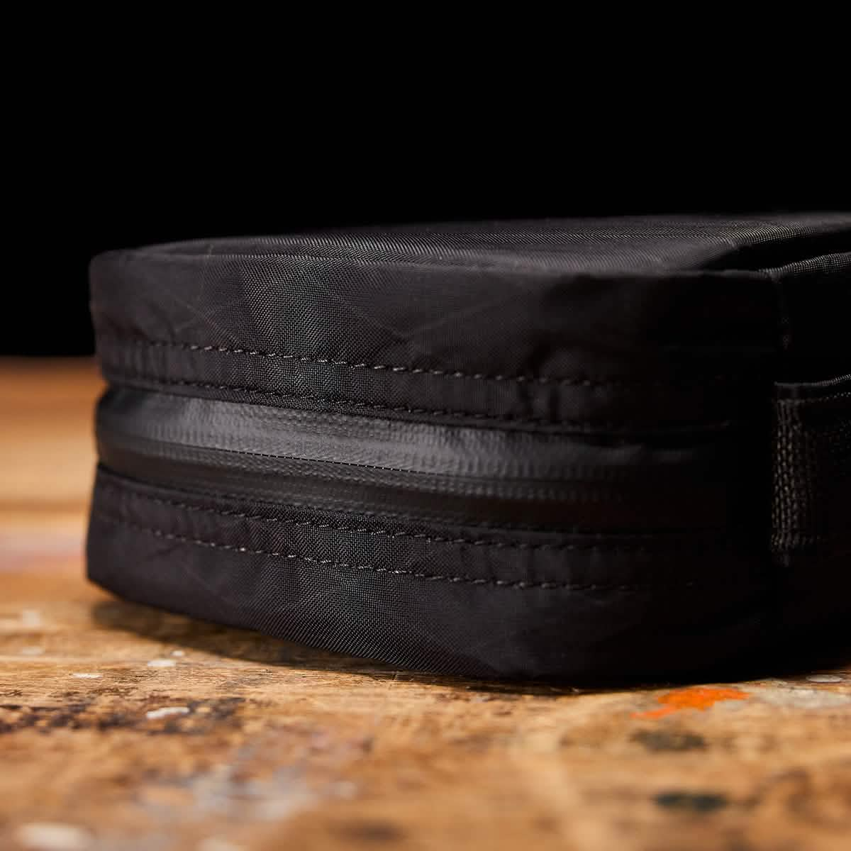 A close-up of the GORUCK GR1 Field Pocket USA - X-PAC, a sleek black rectangular travel accessory expertly crafted with X-PAC fabric and a zipper, resting on a wooden surface. The dark background highlights its texture and precise stitching details.