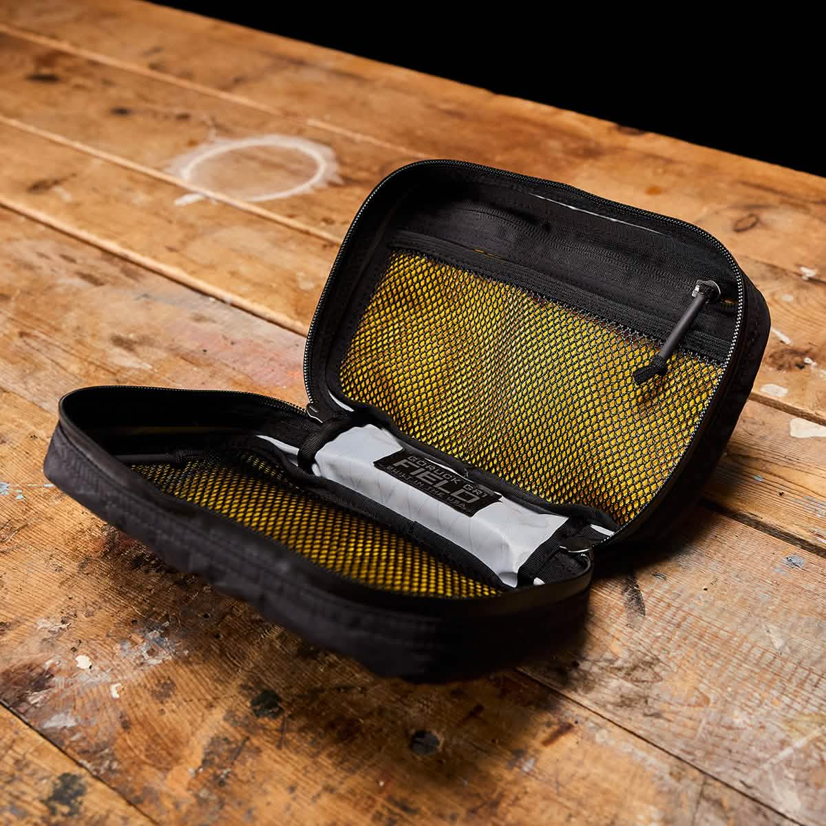 A black zippered storage pouch from GORUCK, similar to the GR1 Field Pocket USA - X-PAC, sits atop a rustic wooden surface. Its yellow mesh pockets stand out against the dark background, emphasizing this X-PAC travel accessory's compartments.