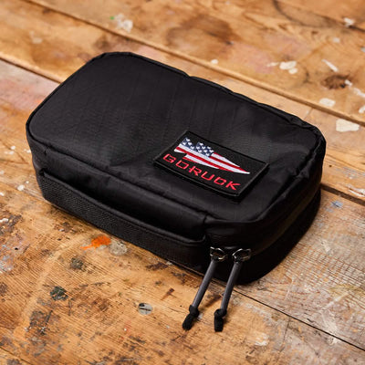 A GR1 Field Pocket USA - X-PAC by GORUCK, adorned with an American flag patch and ideal for travel, rests on a wooden surface. Its zippered closures with two pull tabs provide an elegant contrast to the textured wood and visible paint splatters.