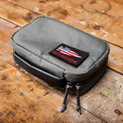 The GR1 Field Pocket USA - X-PAC by GORUCK, an ideal travel accessory, features an American flag patch on top and dual zippers with gray pull cords. It is elegantly displayed on a rustic wooden surface, highlighting its durable X-PAC construction.