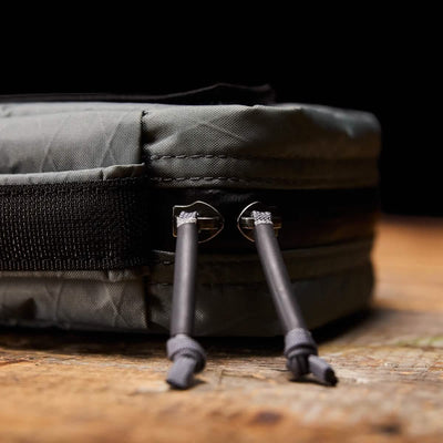 A close-up of the GR1 Field Pocket USA - X-PAC by GORUCK, a versatile travel accessory in black, showcases two zippers with cylindrical pulls resting on a wooden surface. Made from durable X-PAC material, this bag boasts reinforced stitching and a textured finish.