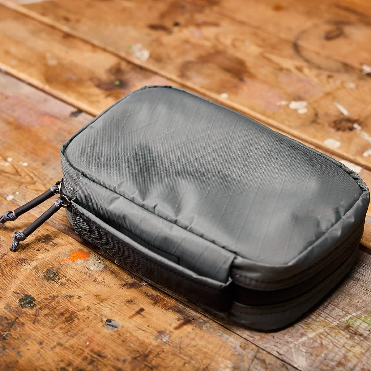 On a rustic wooden surface lies the GR1 Field Pocket USA - X-PAC by GORUCK, a gray rectangular tech pouch constructed from durable X-PAC fabric with zippers. Ideal as a travel accessory, it ensures your essentials stay organized and secure while you're on the go.