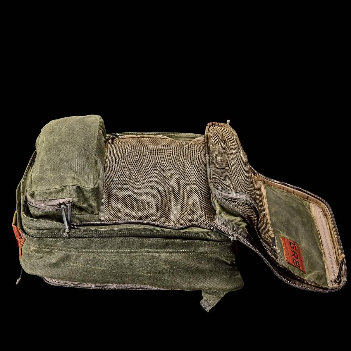 The GR2 - Waxed Canvas by GORUCK, crafted from heritage green waxed duck canvas, stands open to reveal its interior mesh pocket. It features an organizer panel with multiple smaller compartments and is adorned with a brown Red Wing leather tag on the side. The black backdrop enhances its rugged charm.