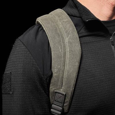 Close-up of a person wearing a black jacket with a GORUCK GR2 - Waxed Canvas rucksack. The green backpack strap, made from waxed duck canvas, rests over their shoulder, highlighting its texture against the jacket.