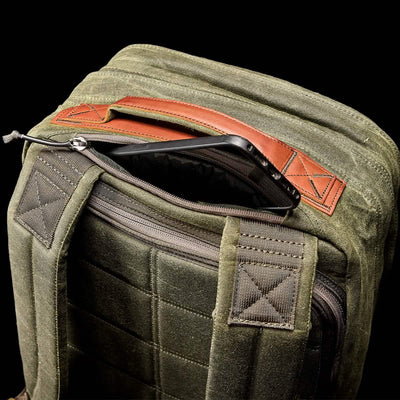 The GR2 - Waxed Canvas by GORUCK is a green backpack made from waxed duck canvas, accented with brown Red Wing Leather. It includes padded shoulder straps and features an open zippered compartment revealing a smartphone against a black background.