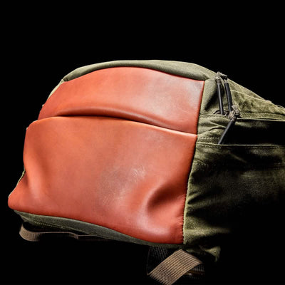 A close-up of the GR2 - Waxed Canvas by GORUCK showcases its exquisite Red Wing Leather front and dark green waxed duck canvas sides. The design features visible zippers and a small external pocket, all set against a black background.