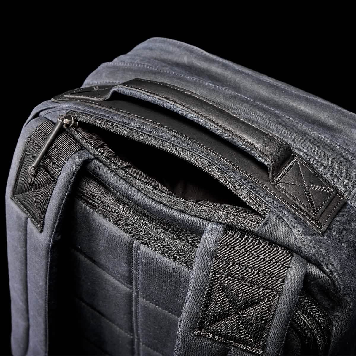 Close-up of a dark grey backpack against a black background. The GORUCK GR2 - Waxed Canvas features padded shoulder straps, a zippered main compartment, and a sturdy handle. Its sleek and modern design showcases detailed stitching with durable fabric, reminiscent of classic styles.