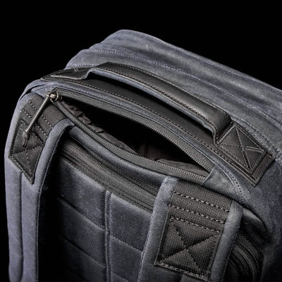 Close-up of a dark grey backpack against a black background. The GORUCK GR2 - Waxed Canvas features padded shoulder straps, a zippered main compartment, and a sturdy handle. Its sleek and modern design showcases detailed stitching with durable fabric, reminiscent of classic styles.