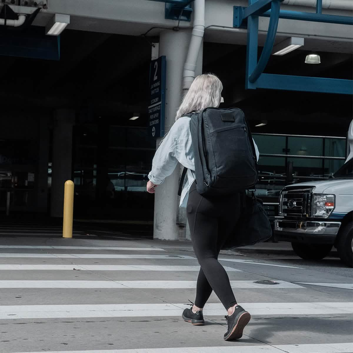 A person with long hair crosses a crosswalk at an airport, carrying a GORUCK GR2 - Waxed Canvas backpack and a duffel bag. They are dressed in black leggings and a light denim jacket. Nearby, a white van is parked under a blue structure.