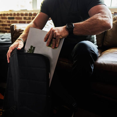 A person is seated on a brown leather couch, placing a laptop adorned with a skateboard sticker into a GR2 - Waxed Canvas rucksack by GORUCK. The scene is warmly lit, with a brick wall in the background.