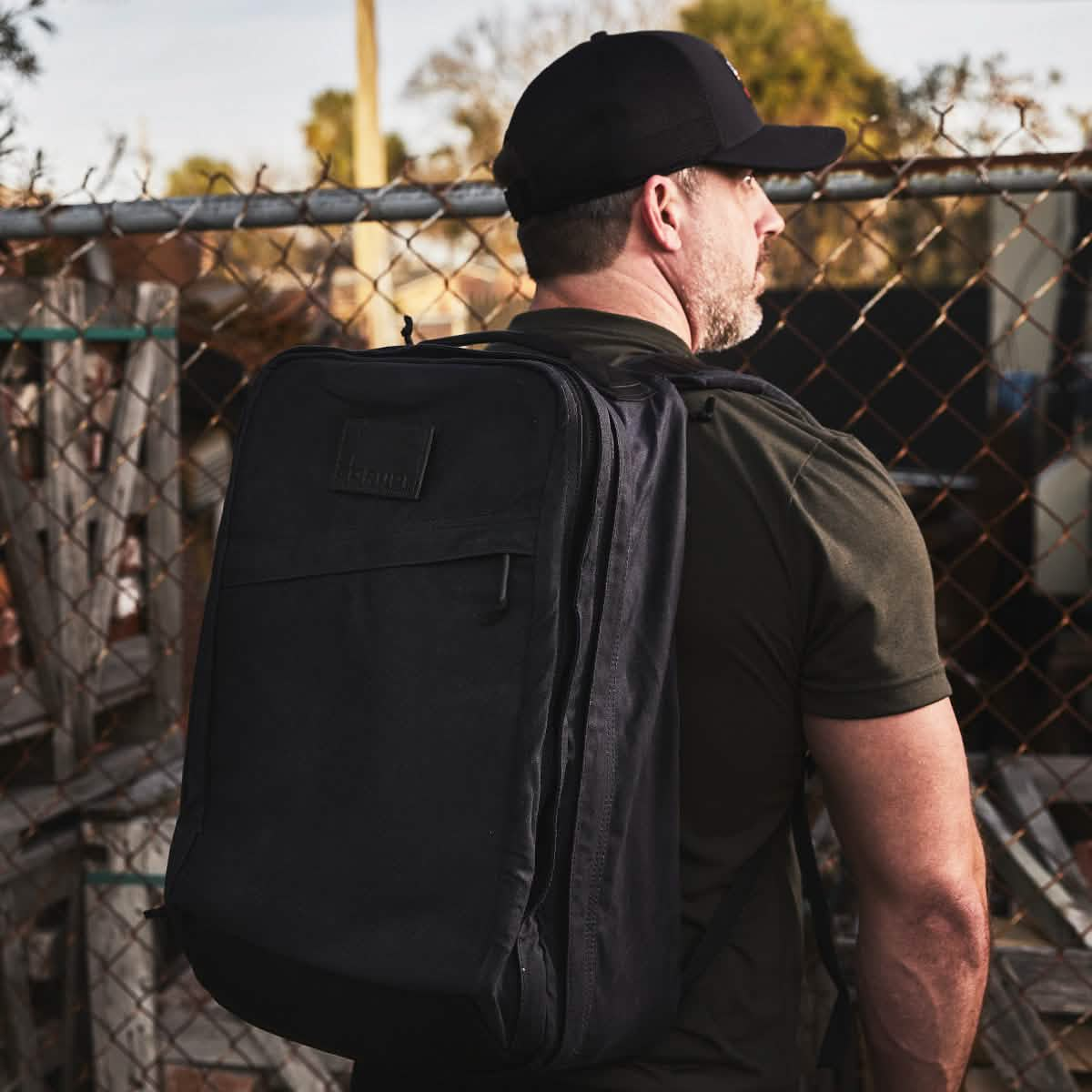 A person wearing a black cap and a large GR2 - Waxed Canvas by GORUCK stands by a chain-link fence. The setting appears to be outdoors with wooden pallets and scattered items in the background.