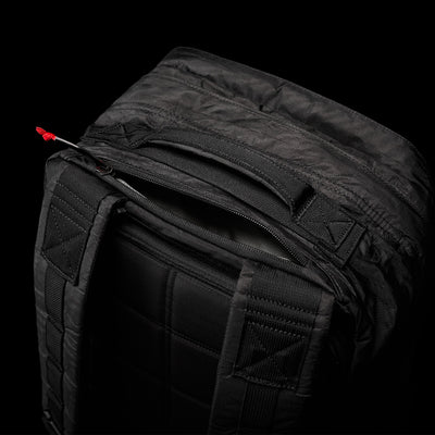 A close-up of the abrasion-resistant black GR2 - Dyneema backpack by GORUCK, with a partially open zippered pocket, reveals padded straps and a quilted back panel. The design features a small red pull tab on the zipper, set against a solid black background.