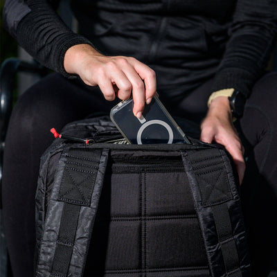 A person dressed in black is slipping a smartphone, protected by a transparent case with a circular design, into an open pocket of the GR2 - Dyneema backpack from GORUCK, known for its abrasion-resistant material.