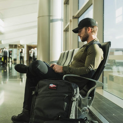 A man wearing a cap and green shirt is seated in an airport terminal, engrossed in his phone. Beside him on the floor is his GR3 - Dyneema carry-on backpack by GORUCK, crafted from durable Dyneema material. Sunlight filters through the large windows, creating shadows where several people can be seen in the background.