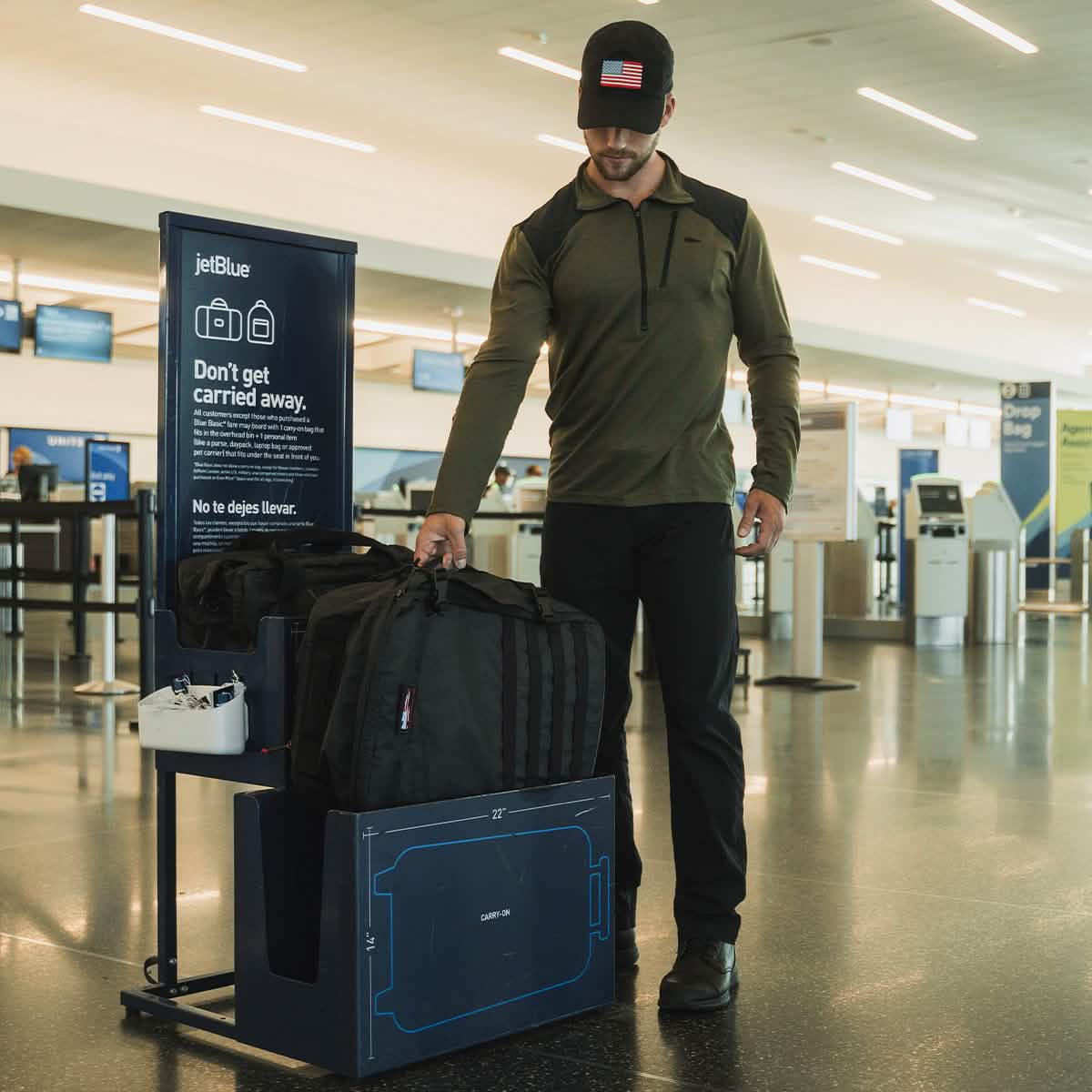 A person in a cap and casual attire is having difficulty fitting their GR3 - Dyneema carry-on backpack by GORUCK into a baggage sizer at an airport. A sign next to them reads, "Don't get carried away," with the airline's name displayed at the top. The airport is lively and bright, with check-in kiosks dispersed throughout.