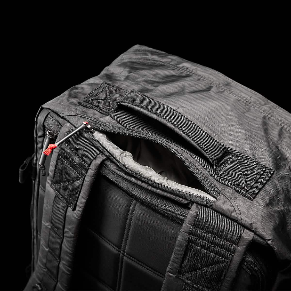 A close-up of the GR3 - Dyneema carry-on backpack by GORUCK sits against a black background, emphasizing the partially open top pocket that offers a peek inside. Made from durable Dyneema material, this backpack comes equipped with padded straps, a handle, and is accented with a distinctive red zipper pull.