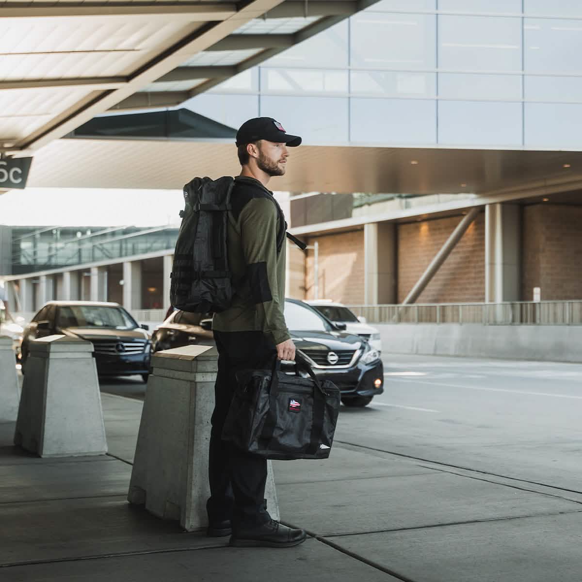 A person stands at an airport curb, facing away. They are dressed in a cap, olive green shirt, and black pants. Over one shoulder is the GR3 - Dyneema carry-on backpack by GORUCK, known for its sleek design and durable material. In their other hand is another bag, while cars are parked behind them under a modern building featuring glass and metal elements.