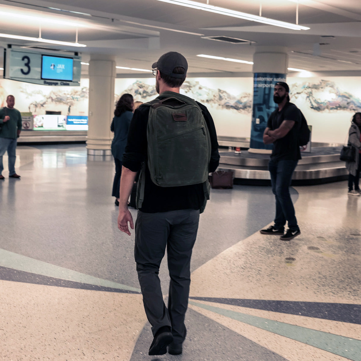 A man wearing a cap and carrying the GR1 USA - Heritage backpack by GORUCK strides through an airport baggage claim area. Other travelers stand around, some retrieving luggage. The space features tiled floors and overhead lighting, with luggage carousels in the background.