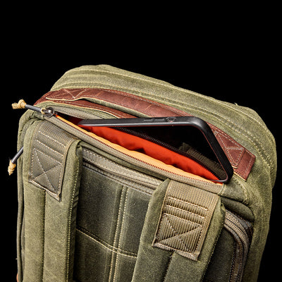 A close-up of the GR1 USA - Heritage by GORUCK in green waxed duck canvas with leather accents. It's partially unzipped to reveal a smartphone inside, with a bright orange interior lining that stands out against the solid black background, highlighting the bag's design and details.