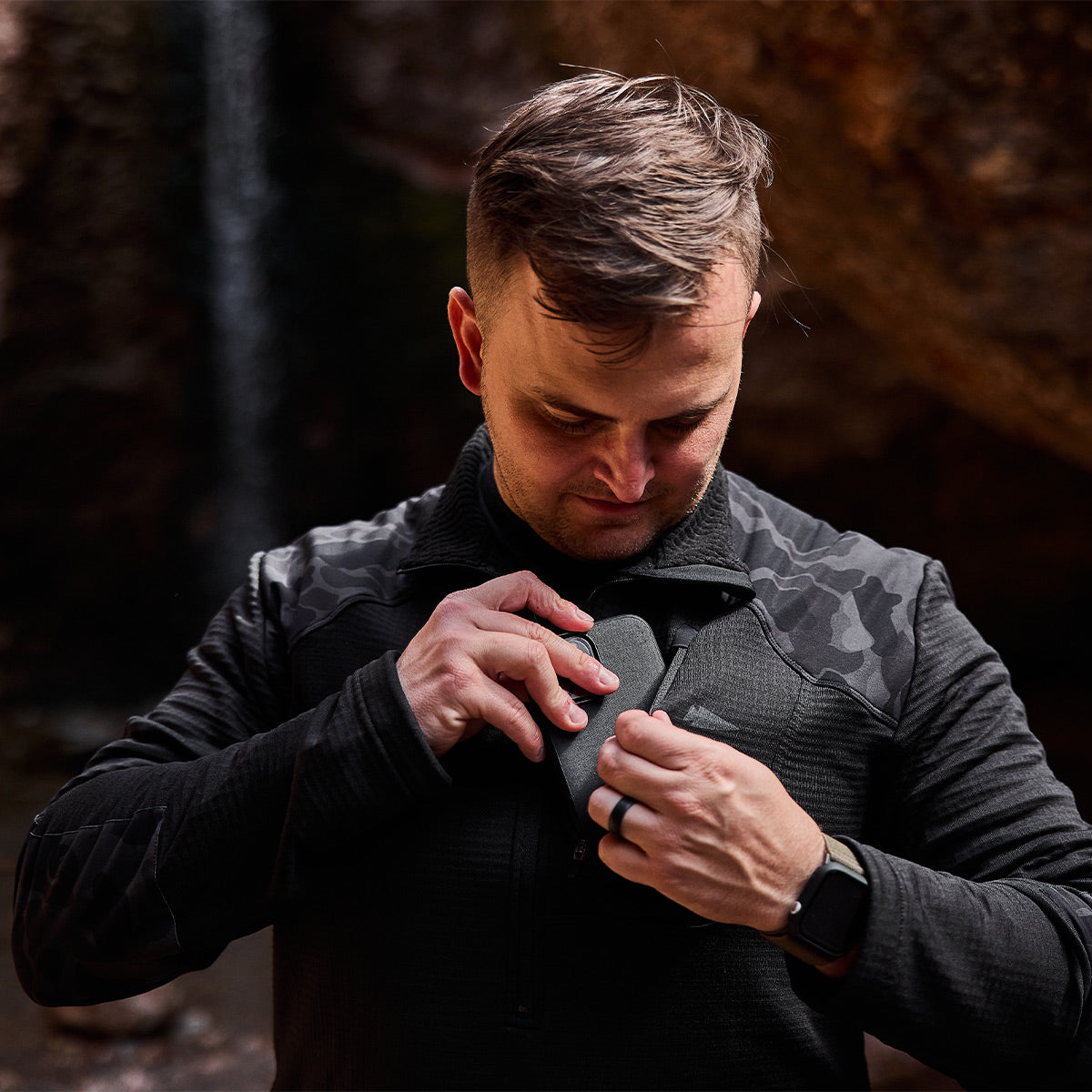 Wearing a Men’s Half Zip - Polartec Grid Fleece, a man stands outdoors, placing an item in his chest pocket while looking down. The rocky backdrop features a serene waterfall cascading behind him.