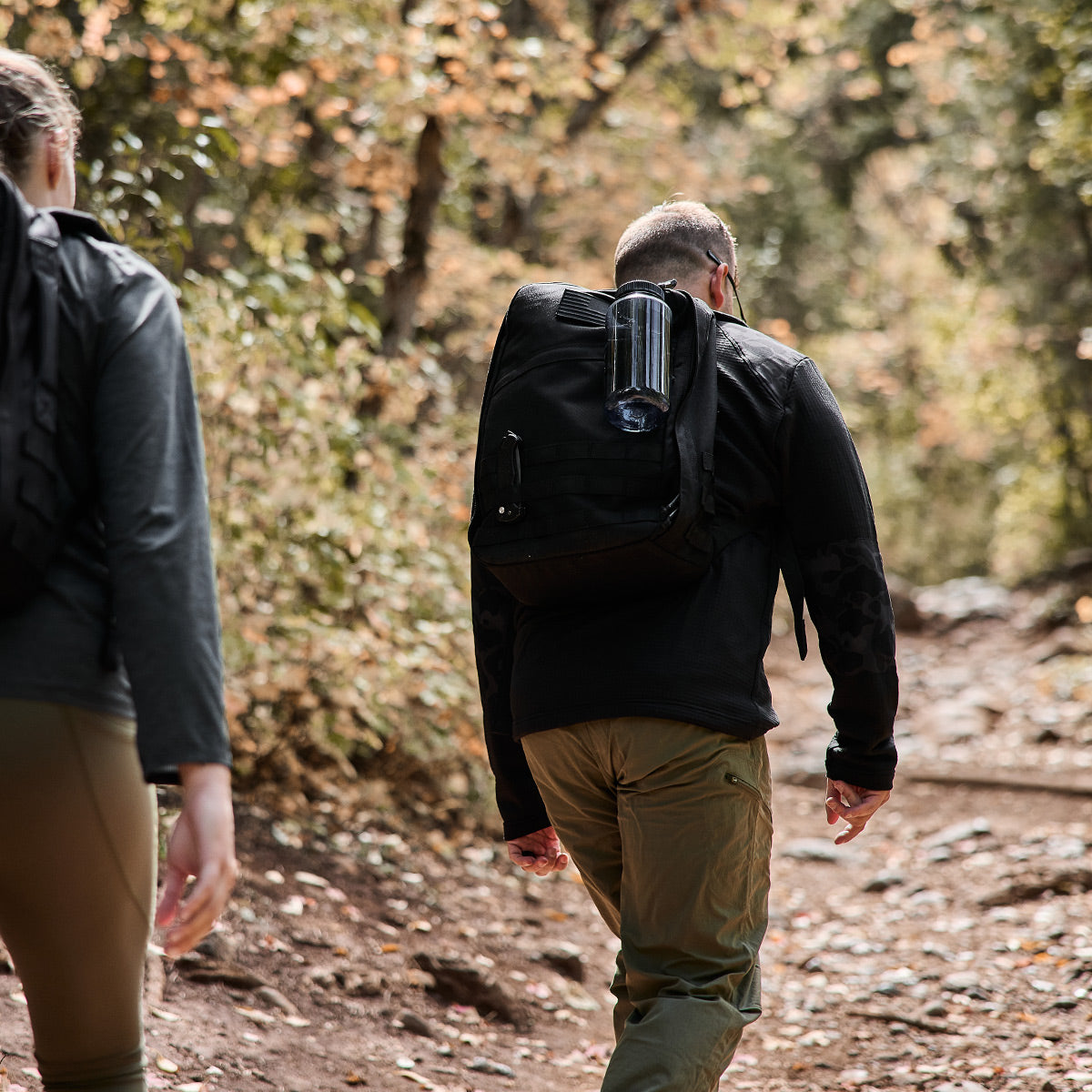 Two people hiking through a sunlit forest wearing Men’s Half Zip - Polartec Grid Fleece, surrounded by green trees and equipped with ToughDry backpacks.