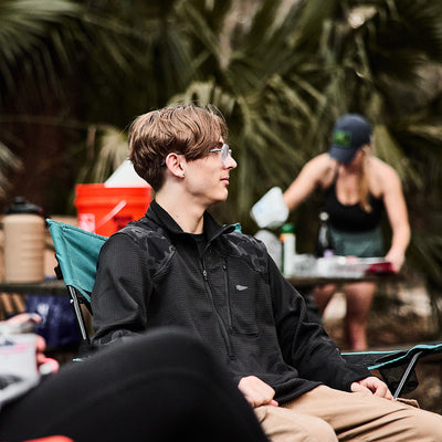 A young man in glasses, wearing a Men’s Half Zip - Polartec Grid Fleece jacket, sits comfortably on a camping chair outdoors; behind him, a woman organizes supplies at the table.