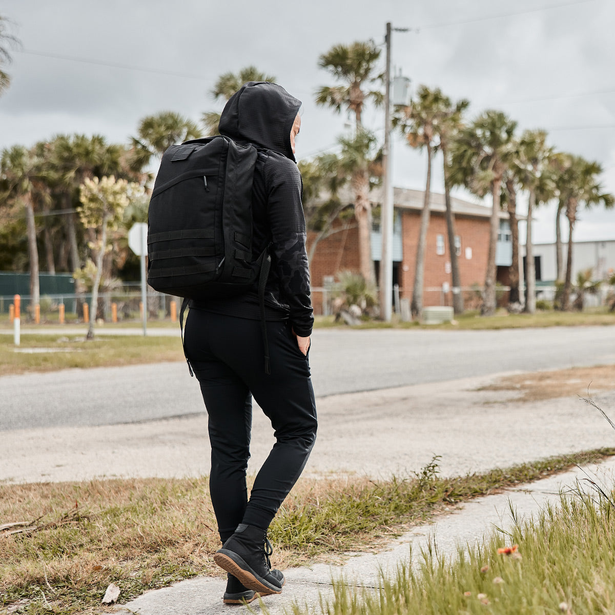 A person wearing the Women’s Full Zip - PolarTec Grid Fleece in black from GORUCK strolls along the sidewalk near the road and palm trees, with their backpack securely fastened.