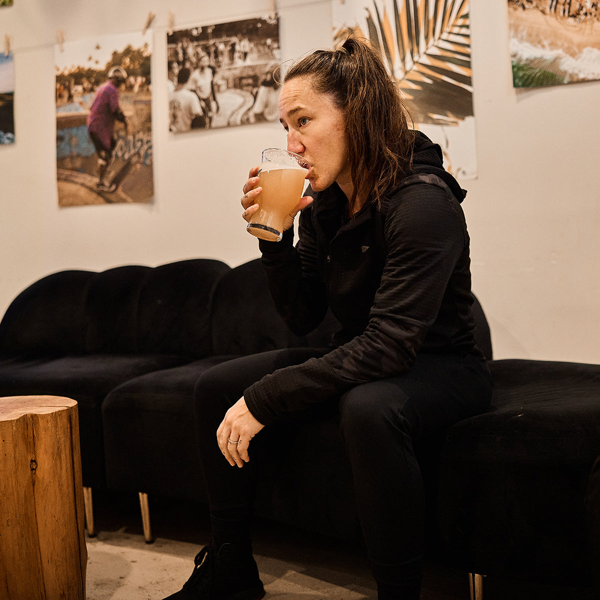 A person sits on a black couch, sipping from a glass in a cozy setting adorned with photos on the wall, wrapped comfortably in GORUCK's Women’s Full Zip - PolarTec Grid Fleece for warmth and efficiency.