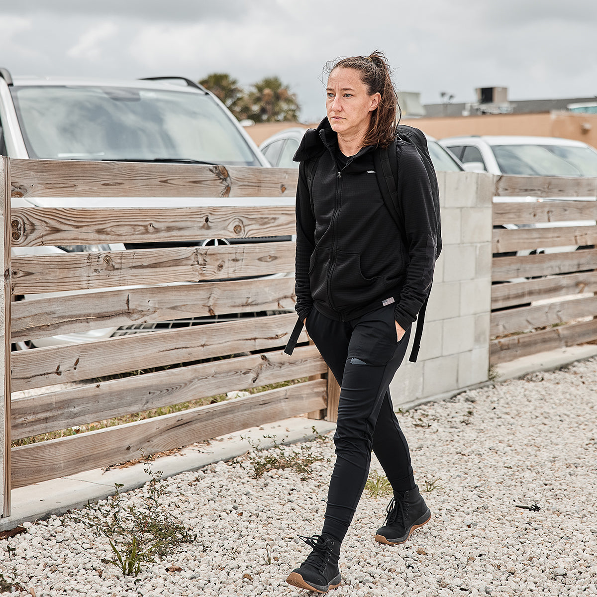 A woman wearing the GORUCK Women’s Full Zip - PolarTec Grid Fleece, in black, strolls along a gravel path near a wooden fence and parked cars.