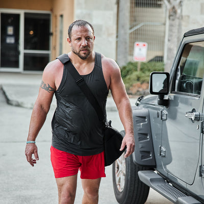 A man dressed in a black tank top and red shorts walks outside near a gray vehicle, carrying a sleek GORUCK Gym Bag made from Ripstop ROBIC®, featuring a shoe compartment. His short hair catches the breeze as he passes by a building entrance and a tree.