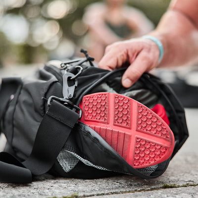 A person puts red-soled sneakers into a black Gym Bag - Ripstop ROBIC® by GORUCK. The bag, showcasing a noticeable buckle and mesh pocket, sits on a stone surface outside. A slightly blurred background suggests an outdoor exercise environment.