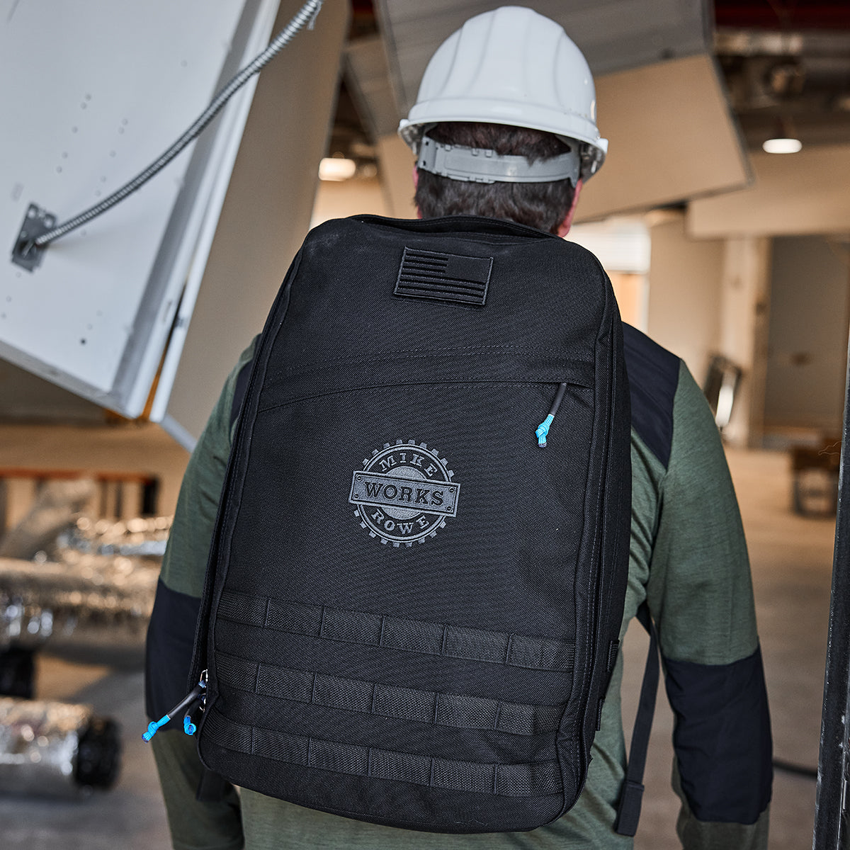 A person stands indoors in a hard hat and green jacket, wearing the GR1 USA GORUCK X mikeroweWORKS backpack. Their gear reflects American craftsmanship, supporting skilled trade initiatives like those by the mikeroweWORKS Foundation.