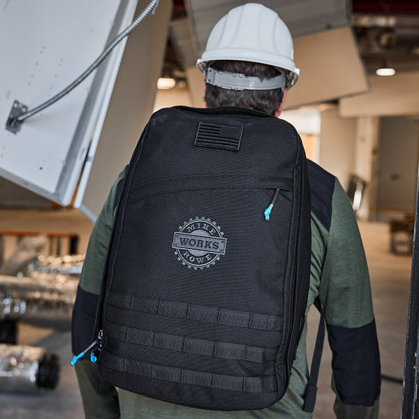 A person stands indoors in a hard hat and green jacket, wearing the GR1 USA GORUCK X mikeroweWORKS backpack. Their gear reflects American craftsmanship, supporting skilled trade initiatives like those by the mikeroweWORKS Foundation.