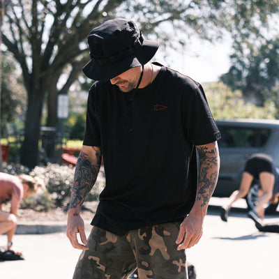 A man wearing a ToughDry Tactical Boonie Hat and a black shirt, with tattoos, stands outdoors while people exercise in the background.