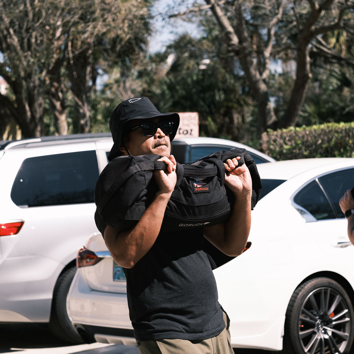 Walking outdoors with a weighted bag on their shoulders, they sport sunglasses and the Boonie Hat - Slick made from ToughDry® fabric, combining style with protection from the elements as cars pass by in the background.