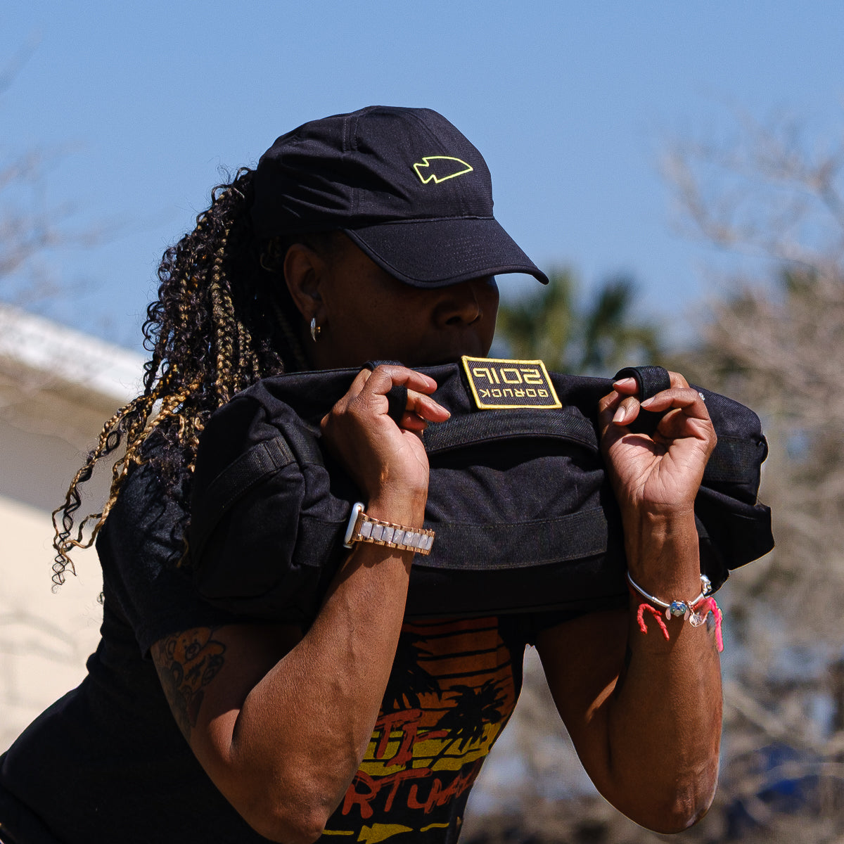 Wearing the Performance Running Hat - ToughMesh, a person showcases strength by lifting a sandbag outdoors, dressed in a graphic t-shirt and bracelets.