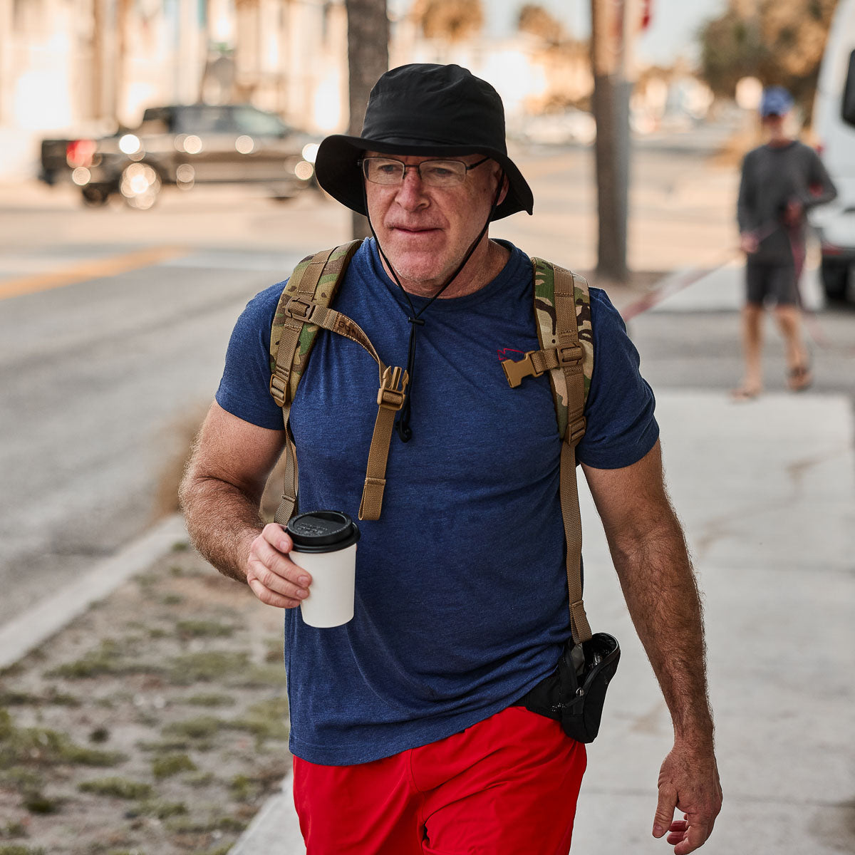 A man in a blue shirt and red shorts strolls the sidewalk, coffee cup in hand. Wearing a black Slick boonie hat made from ToughDry® fabric, he enjoys protection from the elements, with a trusty backpack slung over his shoulder.