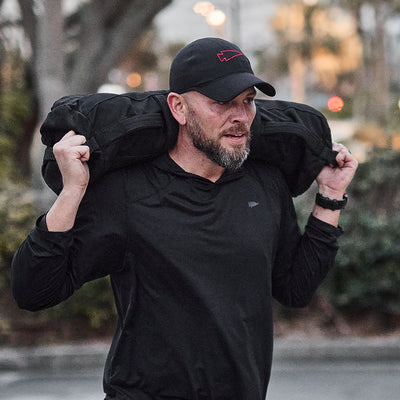 A man in a ToughMesh Performance Running Hat and shirt carries a heavy bag over his shoulders outdoors.