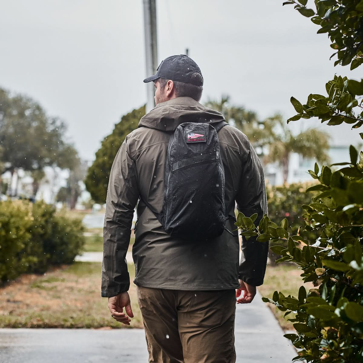 A man in a jacket and cap walks down a wet path, flanked by green bushes and an overcast sky, carrying the Packable Bullet Ruck made from durable Ripstop ROBIC® nylon.